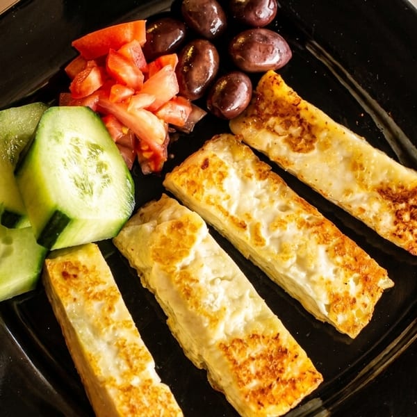 Four golden-brown halloumi cheese sticks with sliced cucumbers, diced tomatoes, and olives on a black plate.