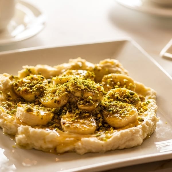A white plate holds a dessert with bananas, cream, honey, and pistachios, with a coffee cup in the background.