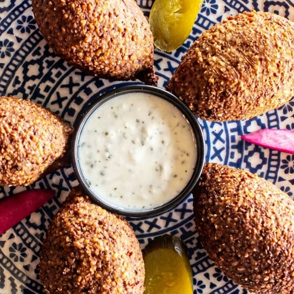 Six kibbeh on a blue and white patterned plate with a side of white sauce and pickles.