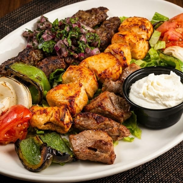 A platter of grilled meats, including chicken, beef, and ground meat, with vegetables and a white dipping sauce.